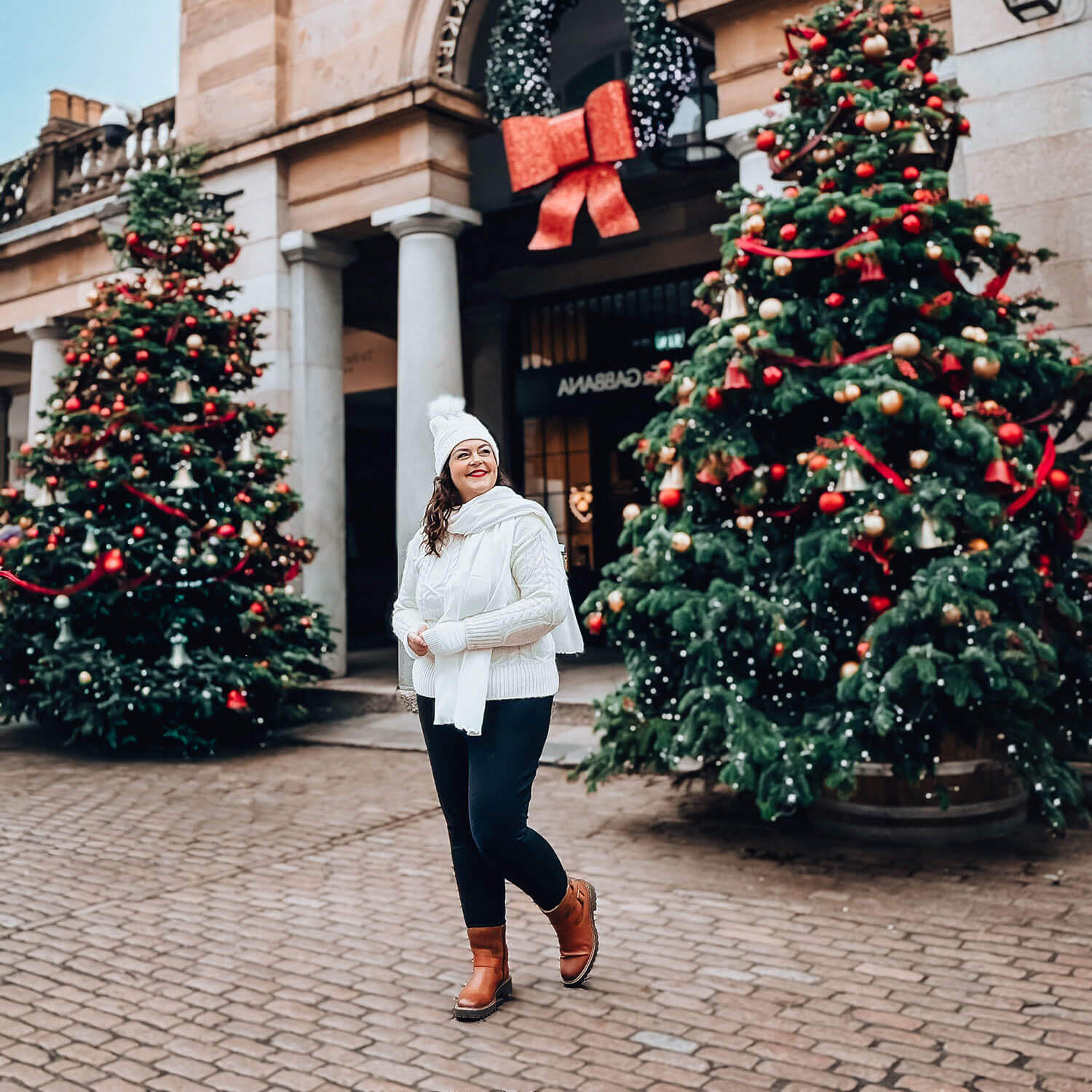 A woman dressed in a white winter outfit walks between two decorated Christmas trees in a festive outdoor shopping area, smiling as she enjoys the scene.
