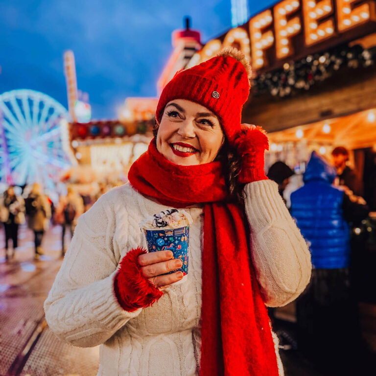 Rosie in a red hat and scarf smiles while holding a festive hot chocolate at a Christmas market, with bright lights and a ferris wheel glowing in the background. The warm, joyful scene captures the atmosphere of Christmas photos.