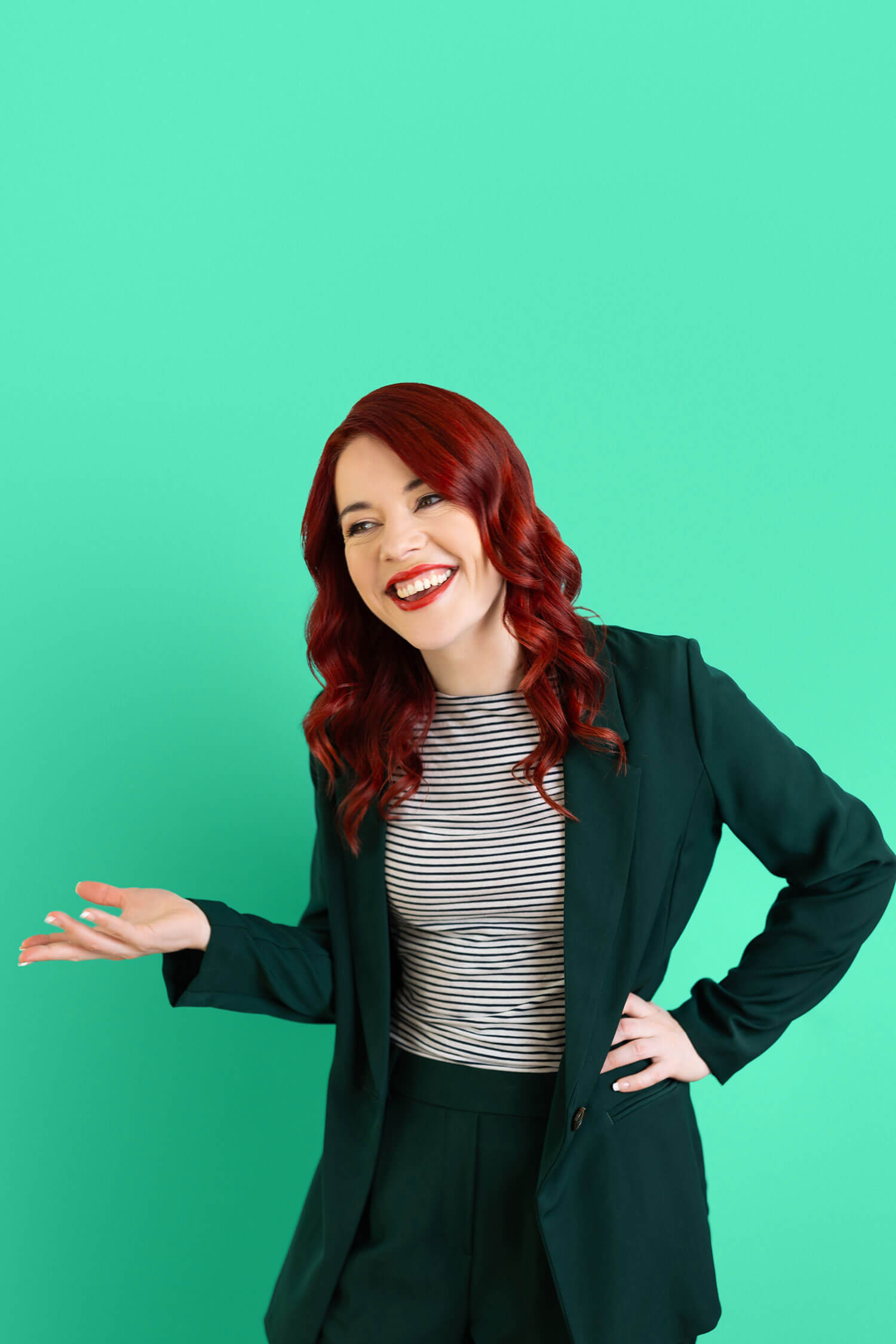 A woman with red hair - Heather Murray from AI for Non Techies - smiles while gesturing with one hand, wearing a striped top and dark blazer against a bright green backdrop during a business photoshoot taken by Rosie Parsons.