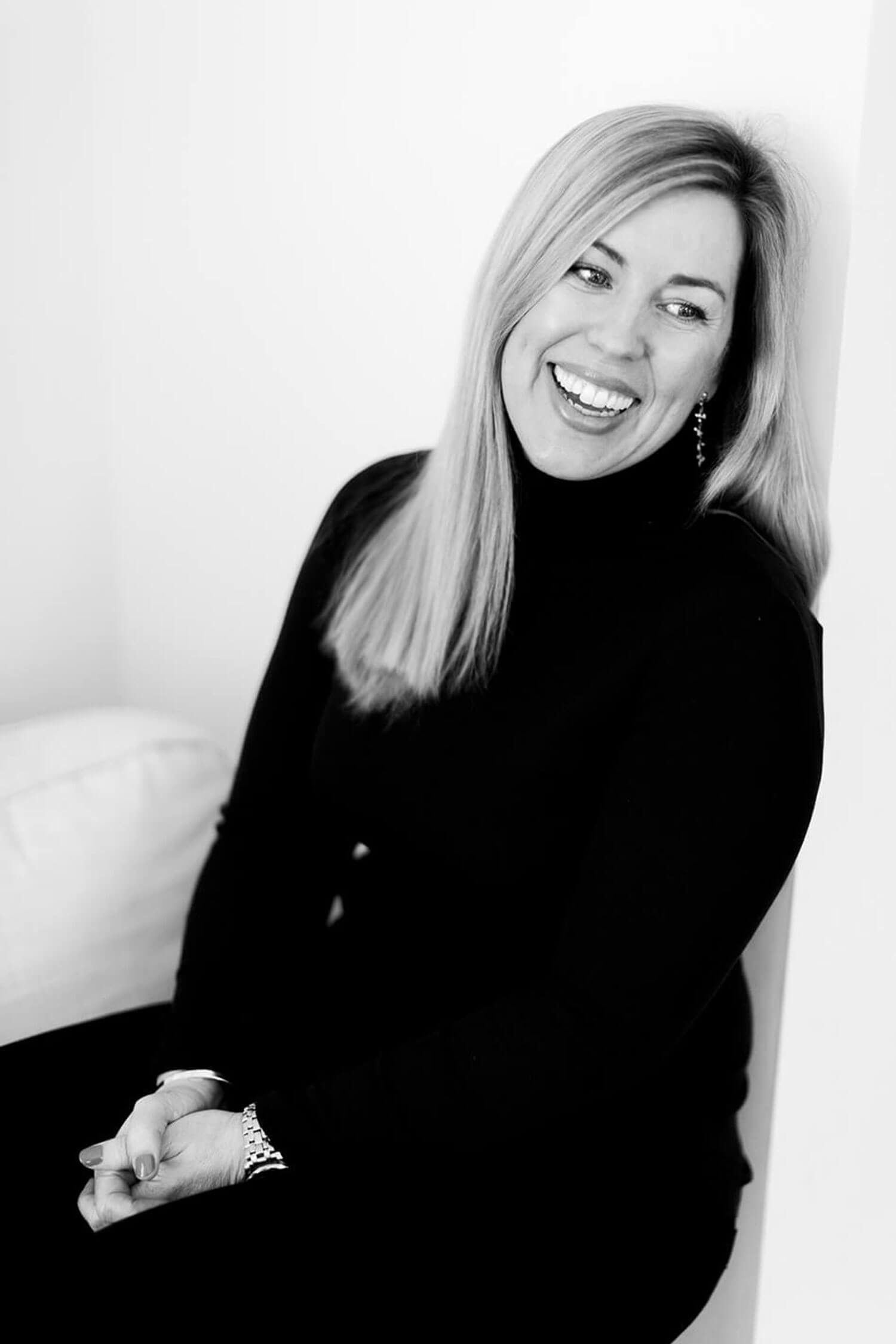 Author Sarah Pearse in a black turtleneck sits with her hands clasped, smiling and looking to the side in a soft black-and-white portrait. Business photoshoot by Rosie Parsons Photography