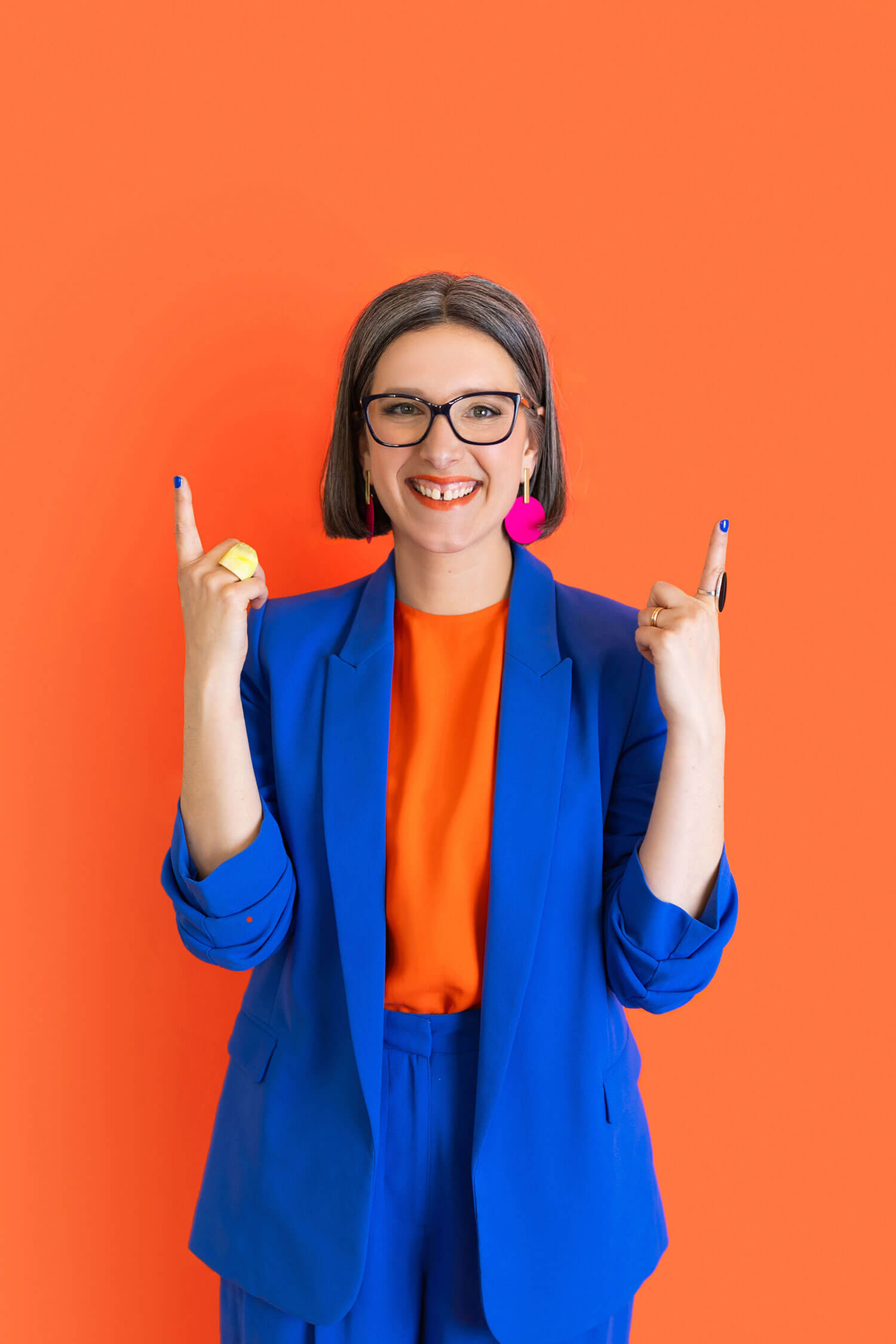 A woman wearing a bright blue suit and orange top smiles and points upwards with both hands against a bold orange backdrop, captured as a colourful professional headshot.