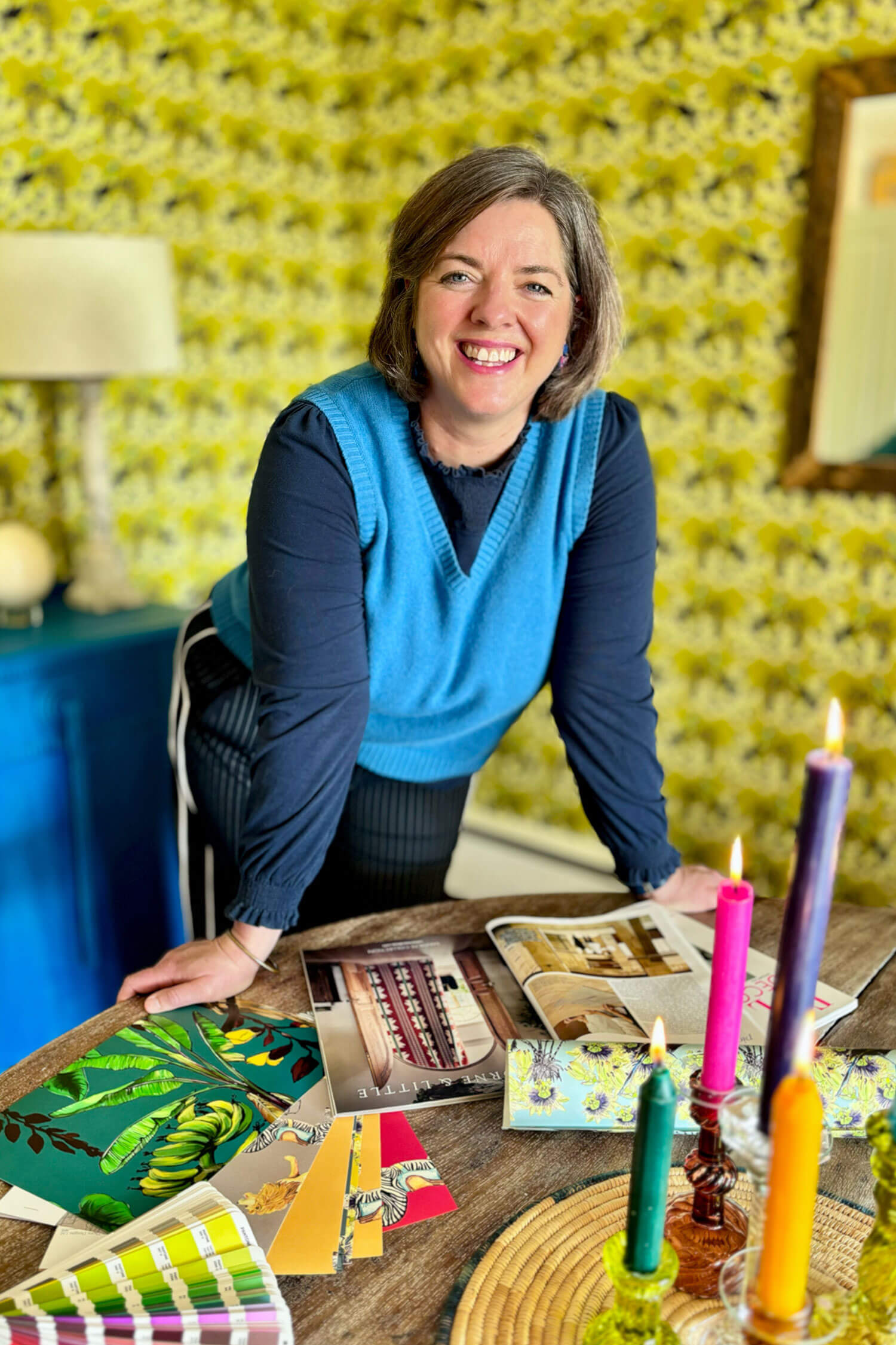 A woman leans over a table covered with colourful wallpaper samples, magazines and candles, smiling in a room with bold yellow patterned wallpaper.