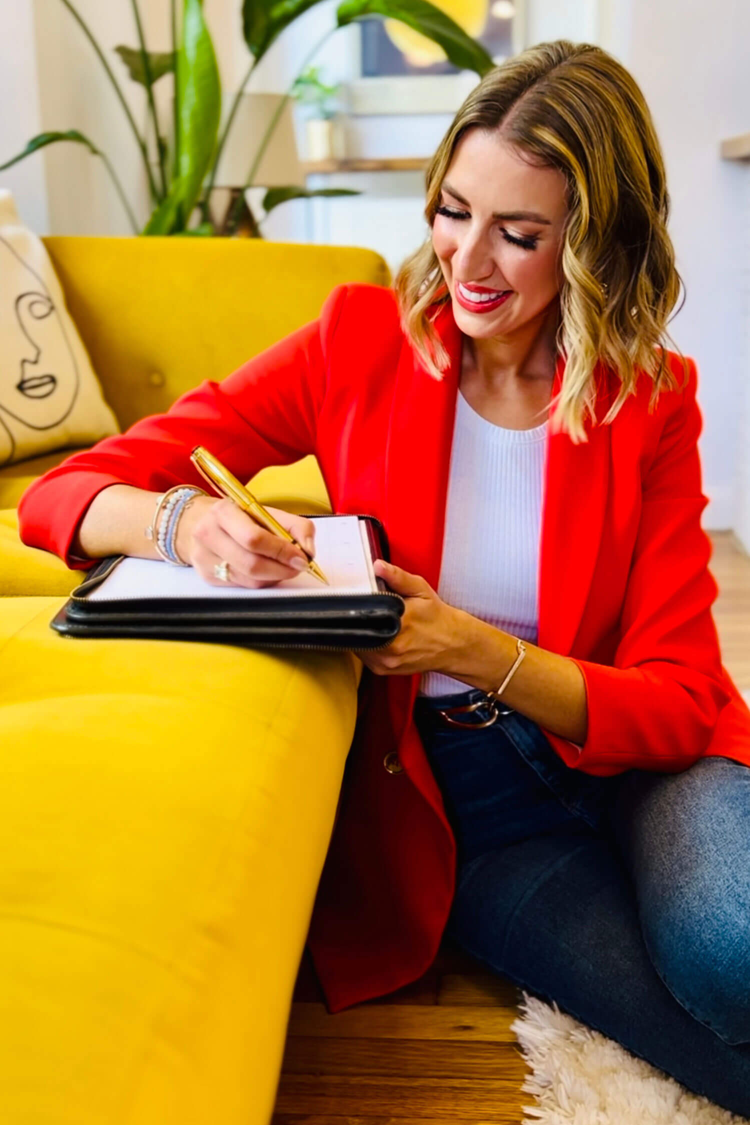 A woman in a bright red blazer sits on the floor beside a yellow sofa, writing in a notebook with a gold pen, captured in a relaxed professional headshot style.