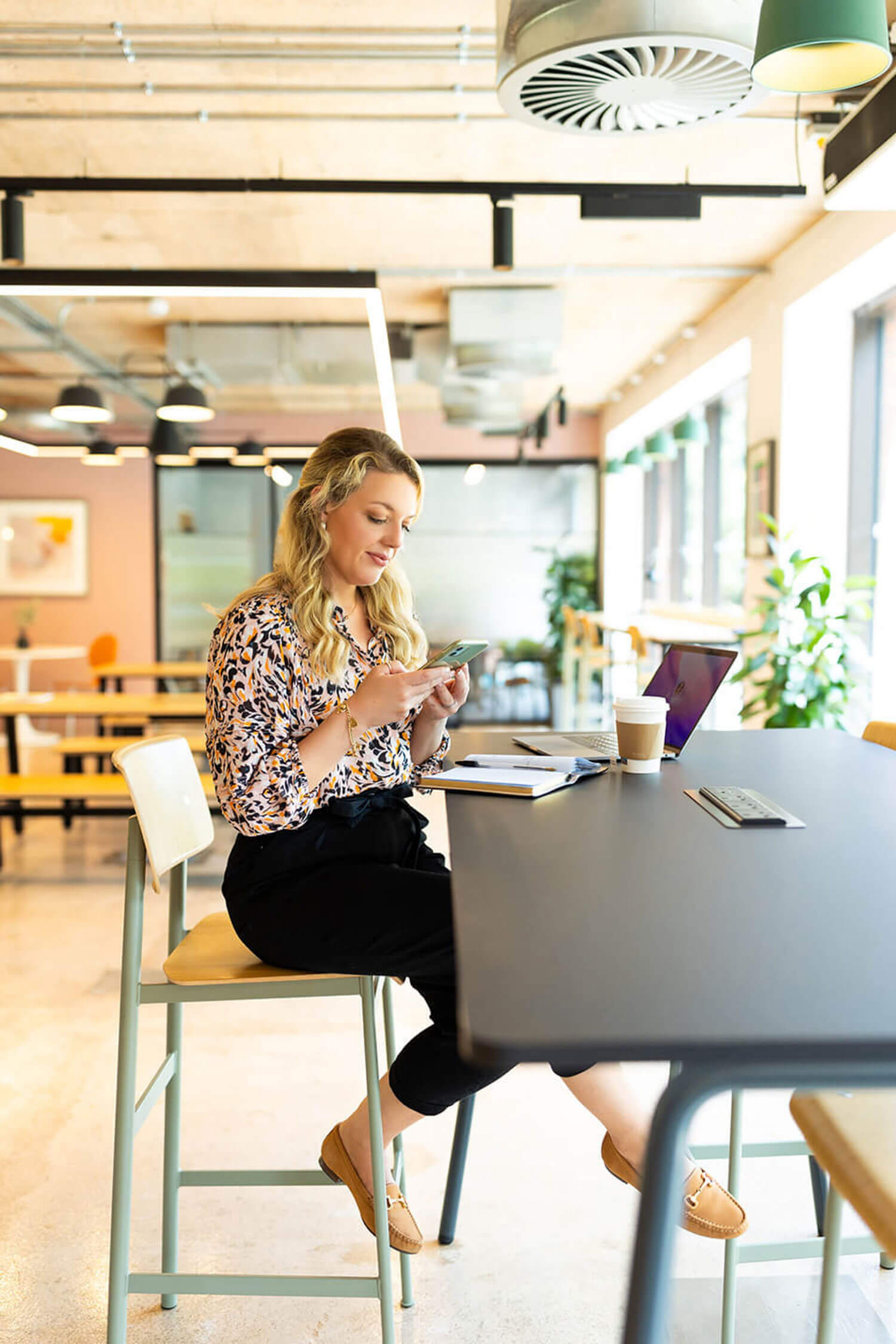 A professional woman with blonde hair checking her phone at a co-working space desk, wearing a leopard print blouse with a laptop and coffee nearby showing inspiration for how to start a blog for beginners.