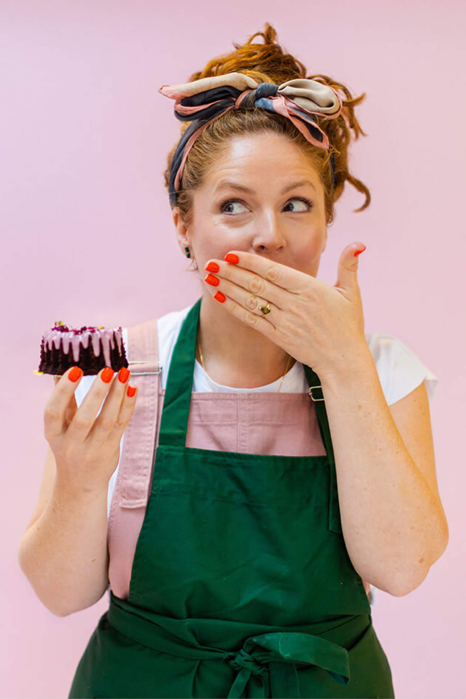 A woman in a green apron holds a small iced cake and covers her mouth with a playful expression, standing against a soft pink background.