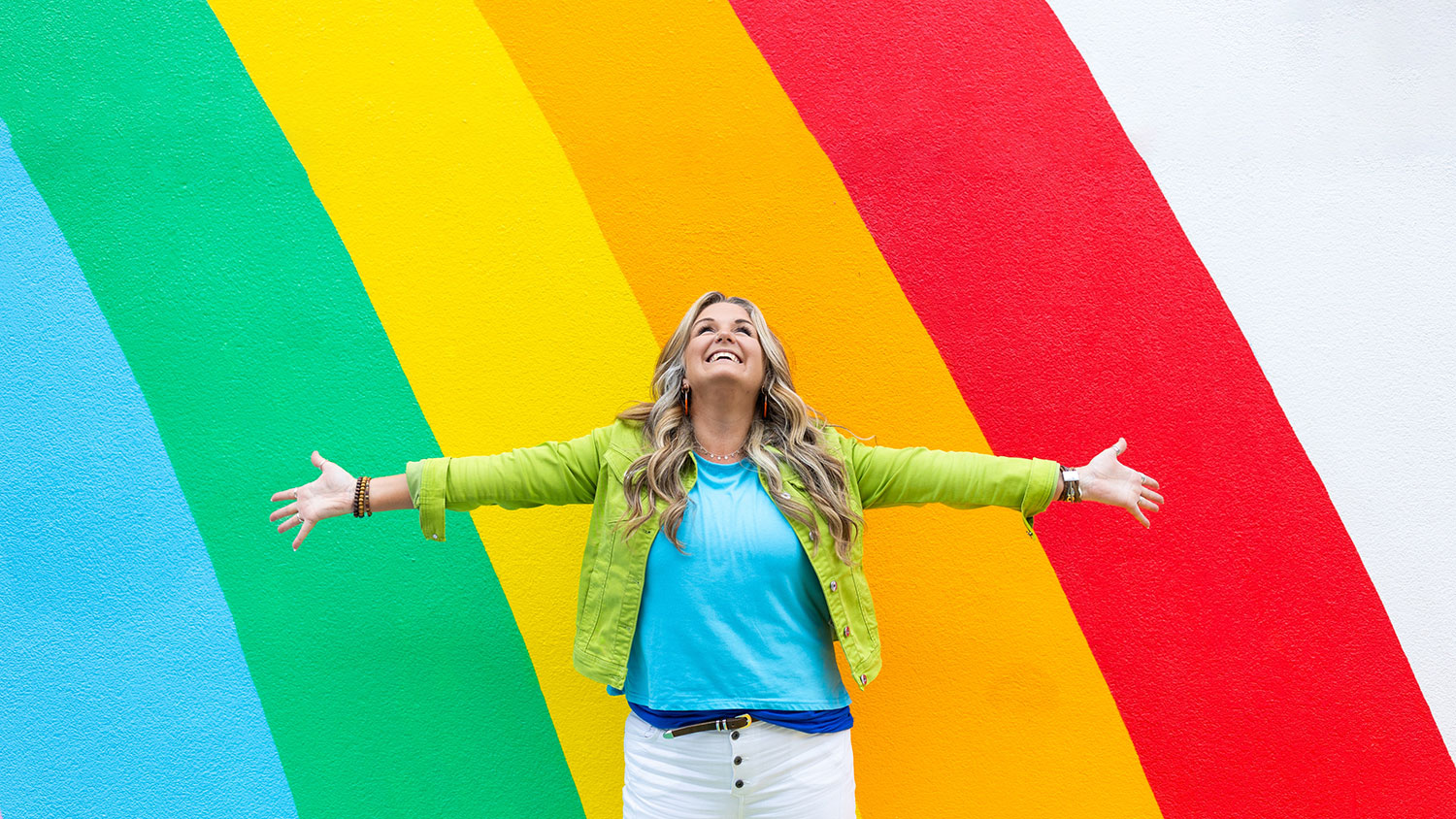 A woman stands in front of a brightly painted rainbow wall with her arms wide open, looking up and smiling, wearing a blue top, white skirt and a green jacket during a business photoshoot.