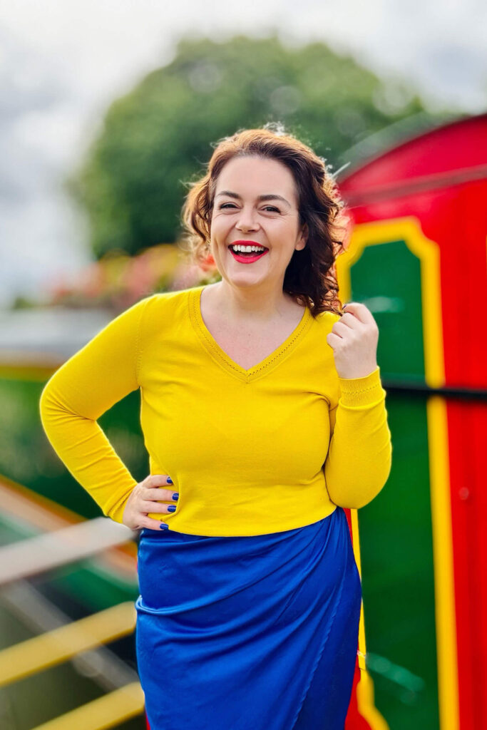 A woman in a bright yellow top and blue skirt laughs outdoors beside a colourful canal boat, captured in a vibrant personal branding portrait that is also a selfie!