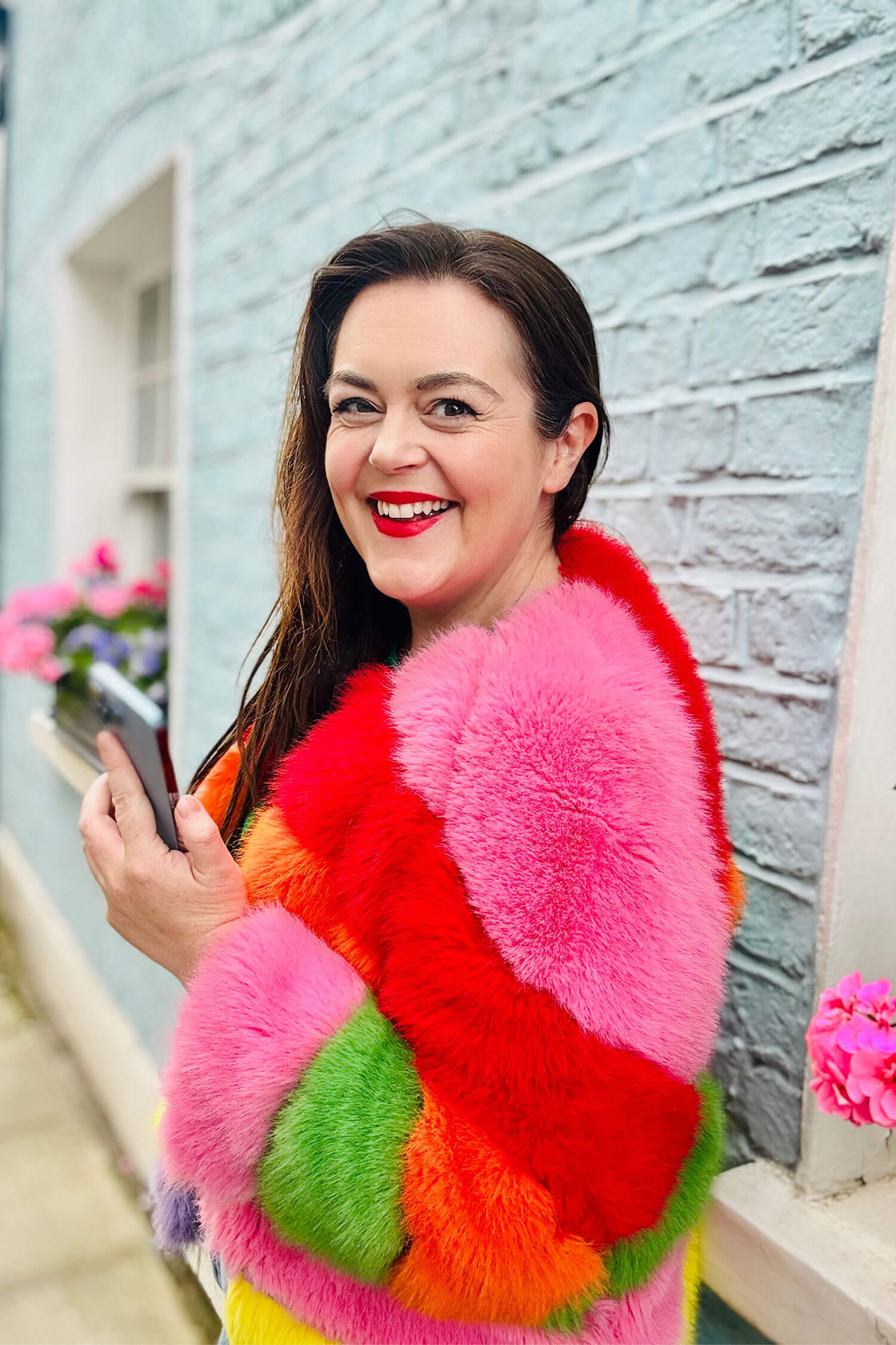 Small business blogger Rosie Parsons smiling and holding a phone, wearing a colourful pink, red, orange and green rainbow fluffy jacket against a pale blue brick wall