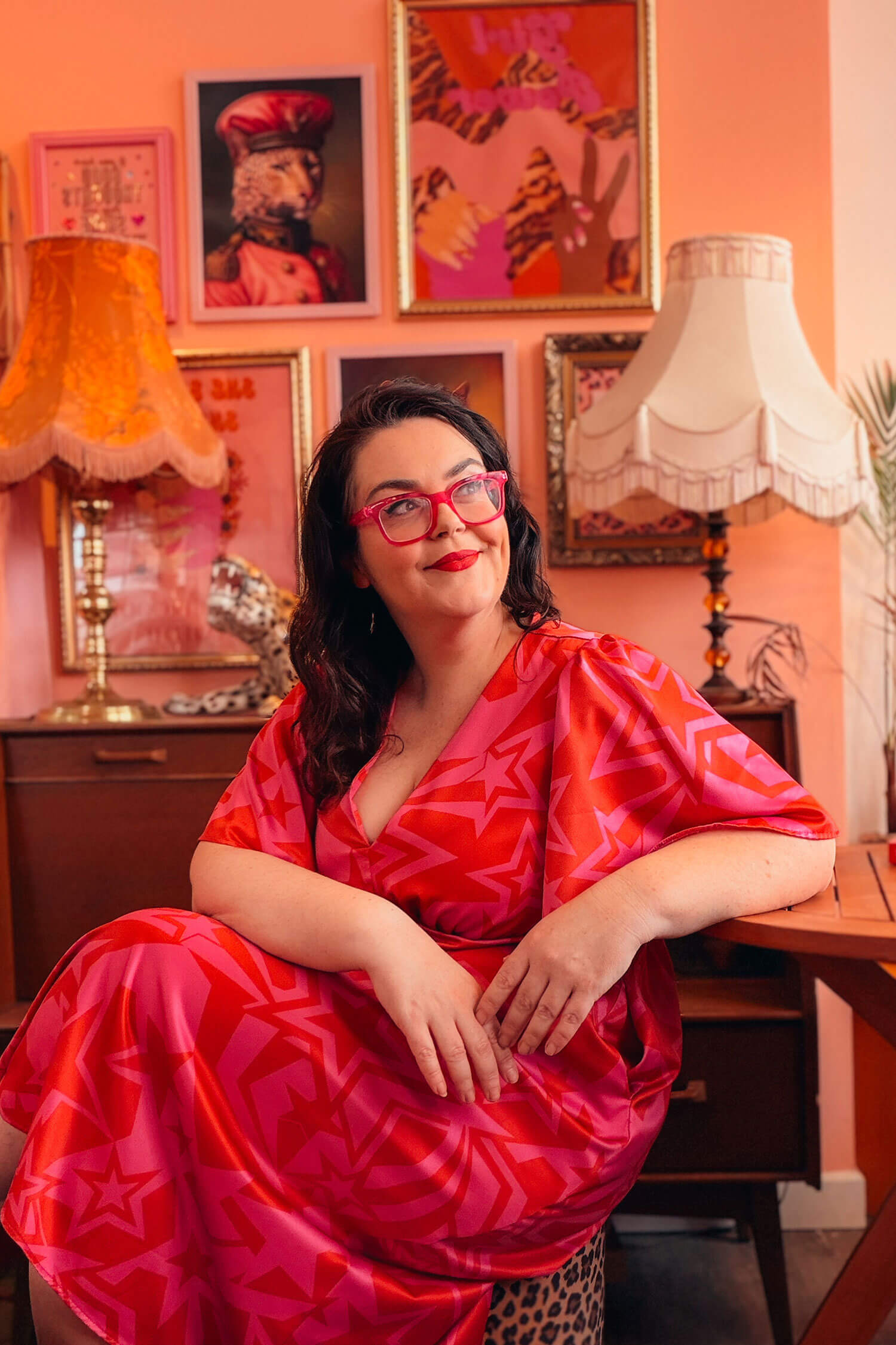 Rosie Parsons in a bold red and pink dress sits in a vintage-inspired room with colourful artwork and lamps, looking off to the side with a relaxed smile during a self portrait business photoshoot.