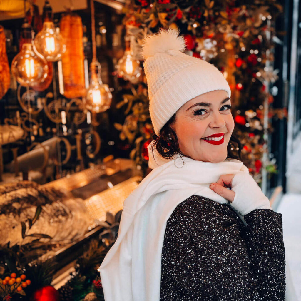 A woman in a white hat and scarf smiles over her shoulder in front of warm Christmas lights and festive decorations during a winter self portrait session.