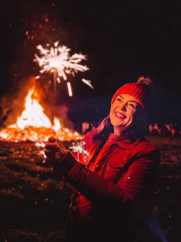 A woman standing outdoors at night smiles while holding a sparkler, with a bonfire and fireworks glowing behind her. The warm autumn colours and festive atmosphere create a cosy seasonal moment that works well for an autumn fall photoshoot
