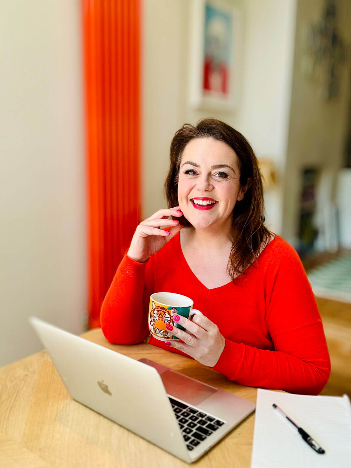 Woman in red jumper sitting at desk with laptop and notepad, holding tiger mug thoughtfully with hand near chin, orange radiator behind, exploring hustles for women