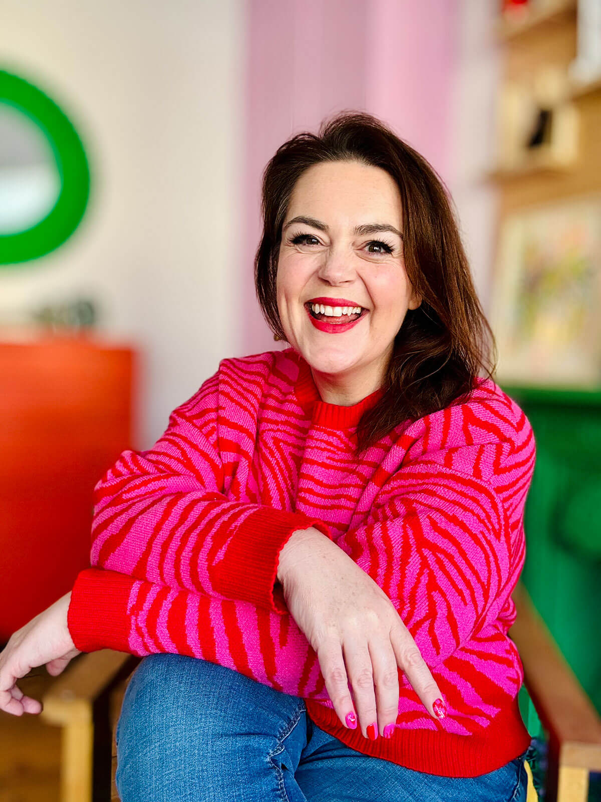 Woman in bold pink and red zebra print jumper sitting relaxed with arms crossed over knee, laughing joyfully in colourful room with green mirror, celebrating work from home jobs