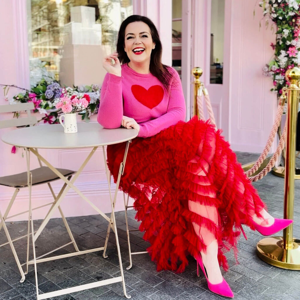 rosie parsons sits outside a pink cafe wearing a pink jumper with red heart and red skirt and pink high heels