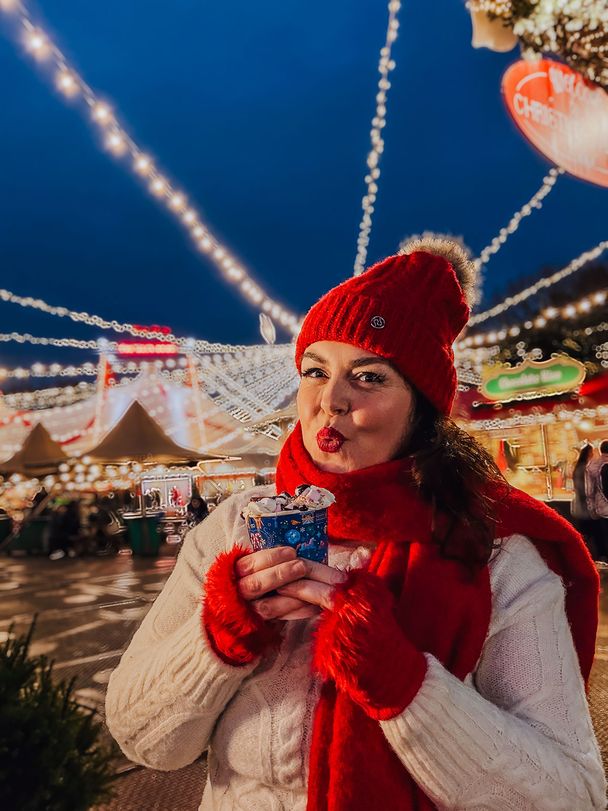 Rosie Parsons, professional photographer and business blogger, blowing a kiss while holding a hot chocolate at a Christmas market during blue hour. She wears a white cable-knit sweater, red beanie, red scarf and red fluffy gloves. Festive string lights and market stalls glow behind her against the deep blue evening sky.