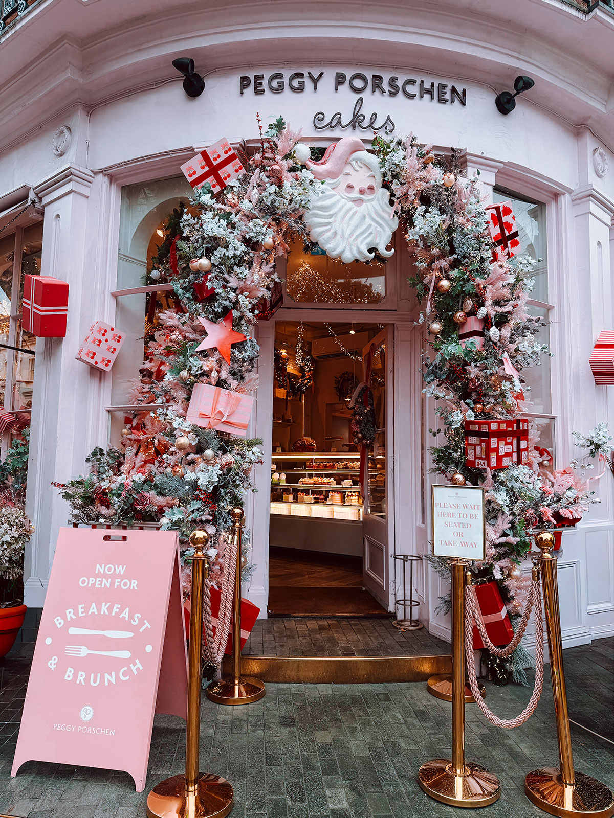 Peggy Porschen Cakes shop front in London decorated for Christmas with pink and white frosted garlands, a Santa face, wrapped gift boxes and baubles framing the entrance. A pink breakfast and brunch sign stands outside with gold rope barriers. The scene shows a creative christmas photoshoot location in London.
