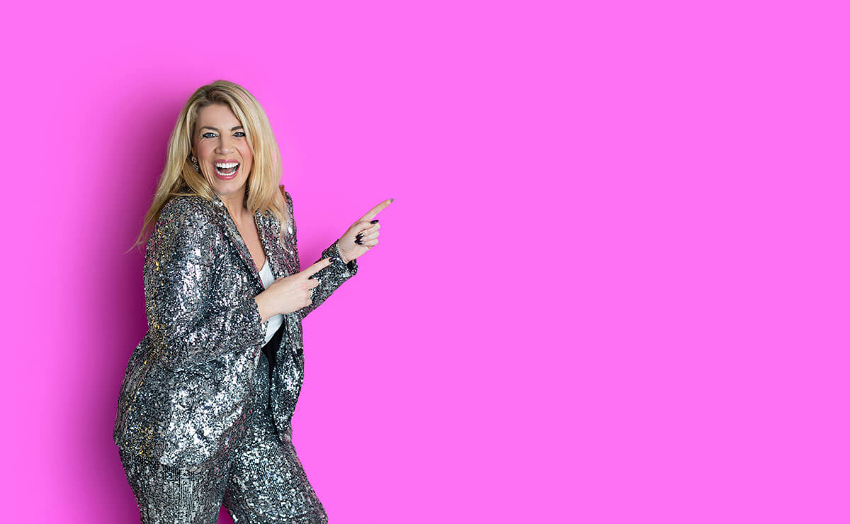 Laura Brunton business coach wears a silver sequin suit while laughing and pointing to the side in a studio personal branding photoshoot against a pink background