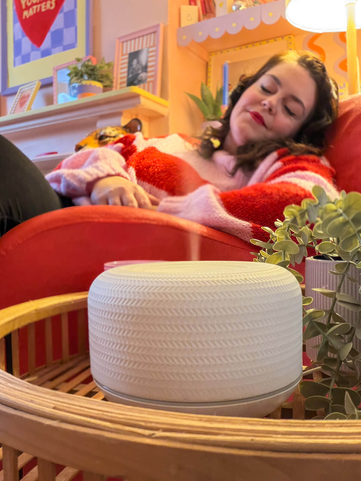 Rosie Parsons, brand photographer and business blogger, relaxing with her eyes closed on a red velvet sofa wearing a pink and red striped jumper. In the foreground is a white textured aromatherapy diffuser on a wooden tray with small potted plants. A cosy, warm lit room with colourful decor in the background. An image representing selfcare ideas, me time and self care day relaxation.