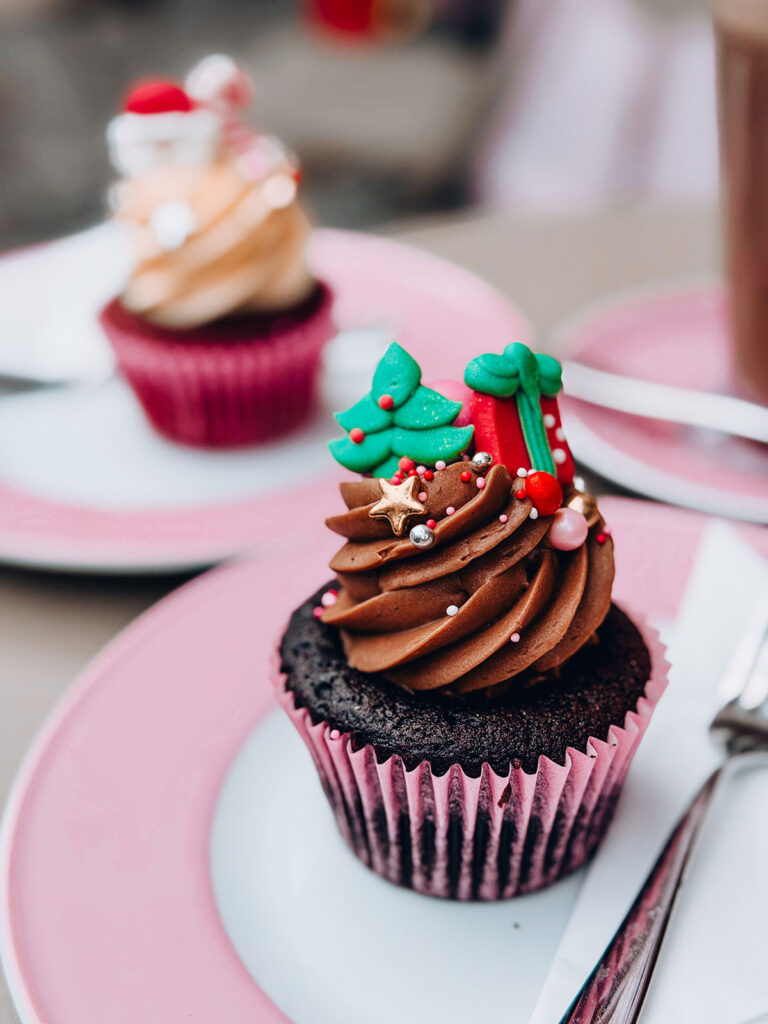 Two festive Christmas cupcakes on pink-rimmed plates. The front cupcake has chocolate frosting topped with a sugar Christmas tree, wrapped gift and gold star decorations. A second cupcake with caramel frosting and a Santa topper sits behind. The scene captures a cosy Christmas aesthetic with warm festive Christmas mood vibes.