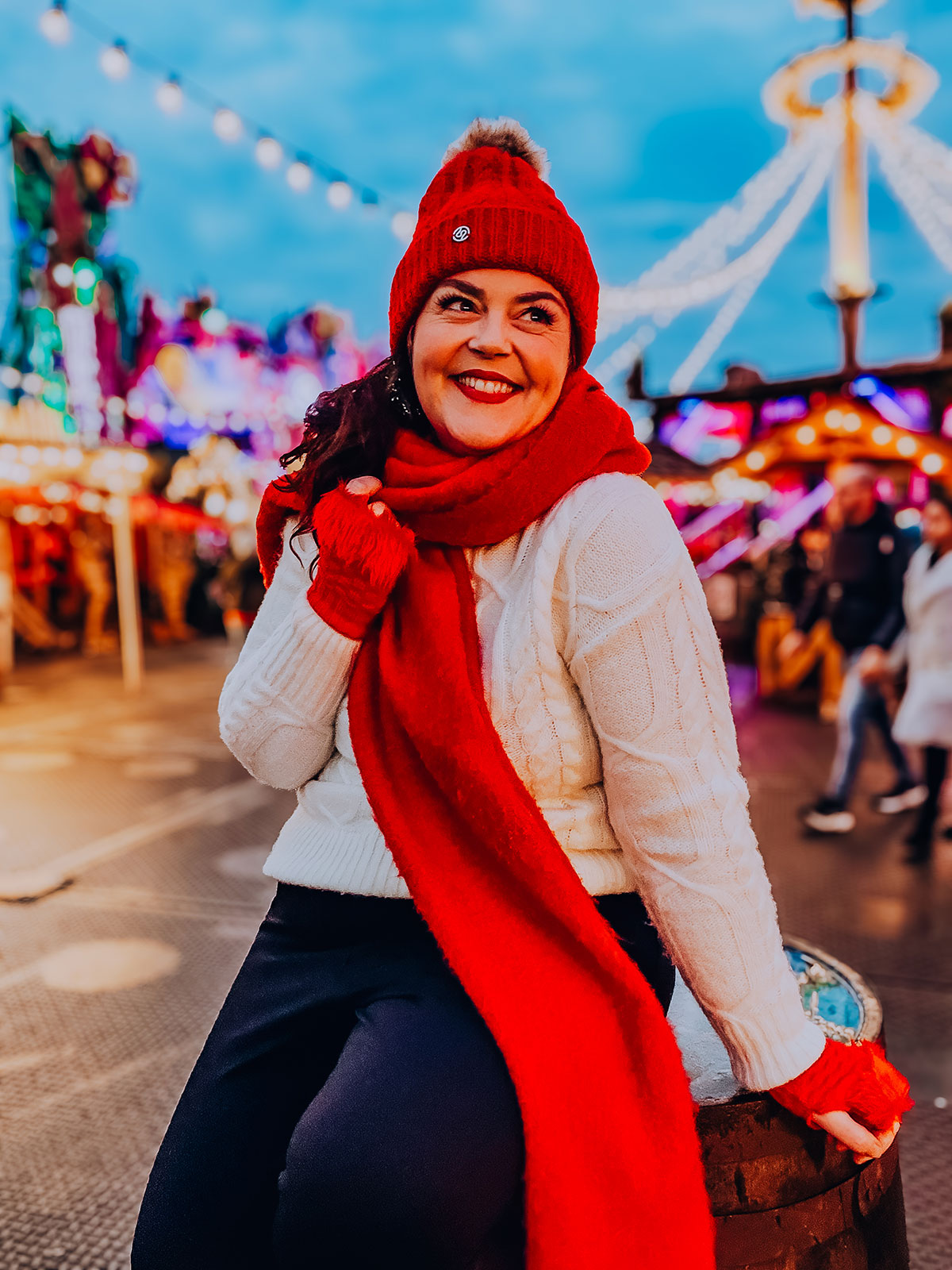 Rosie Parsons, professional photographer and business blogger, smiling at a Christmas market during blue hour. She wears a cream cable-knit sweater, red bobble hat, red scarf and red gloves. Festive string lights and market rides glow against the deep blue evening sky, showing christmas poses at dusk.