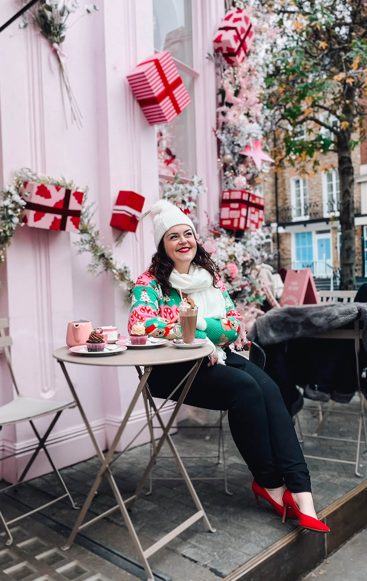 Rosie Parsons, professional photographer and business blogger, sitting outside a pink decorated cafe holding a hot chocolate. She wears a green Christmas jumper with festive print, white bobble hat, white scarf and red heels. Cupcakes sit on the table with pink wrapped gift boxes on the wall behind, showing a creative christmas photoshoot setup.