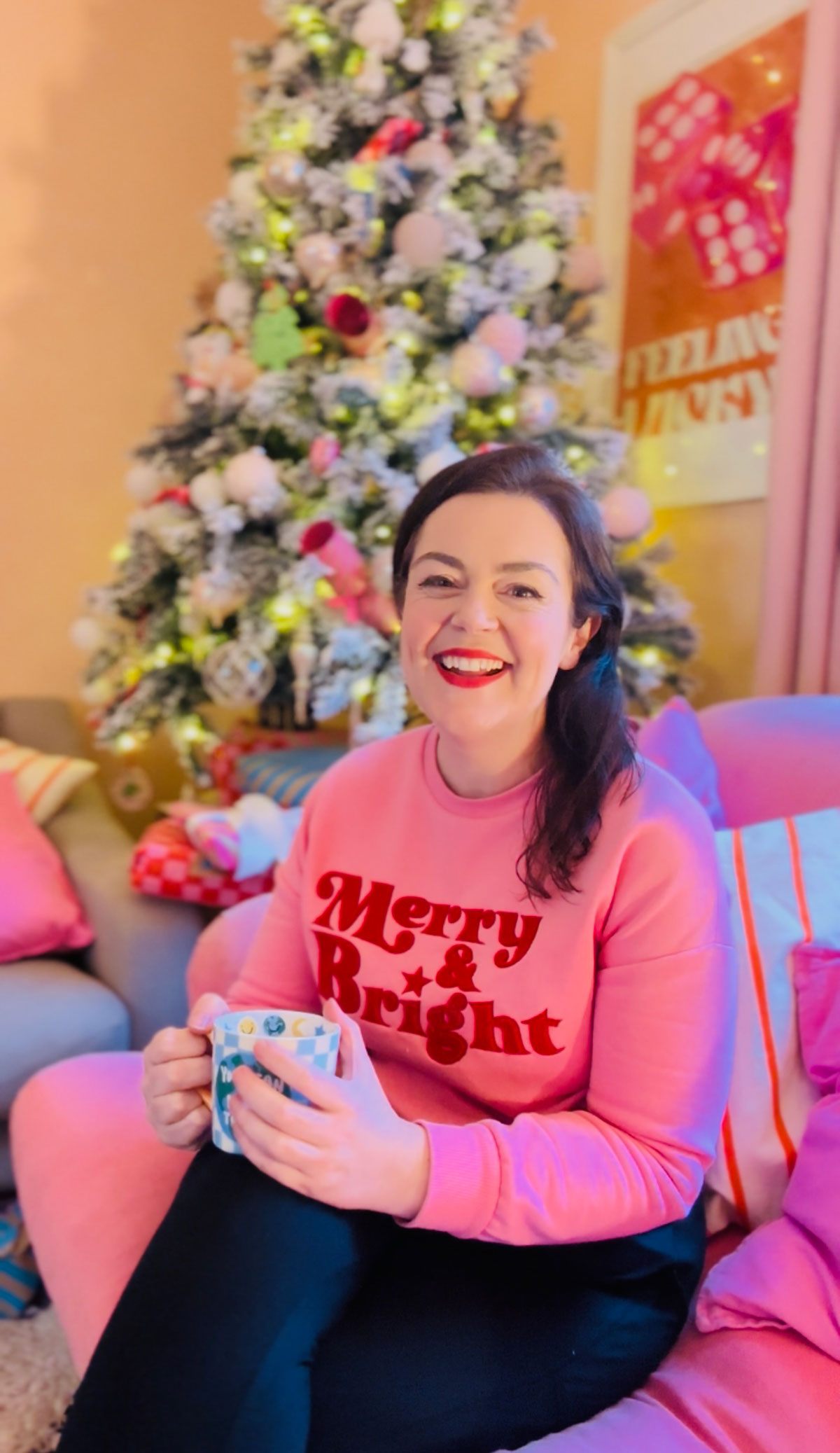 Rosie Parsons, professional photographer and business blogger, laughing while holding a mug on a colourful sofa. She wears a pink "Merry & Bright" sweatshirt with a frosted Christmas tree decorated with pink baubles behind her. The relaxed scene shows a cozy christmas photoshoot setup at home.