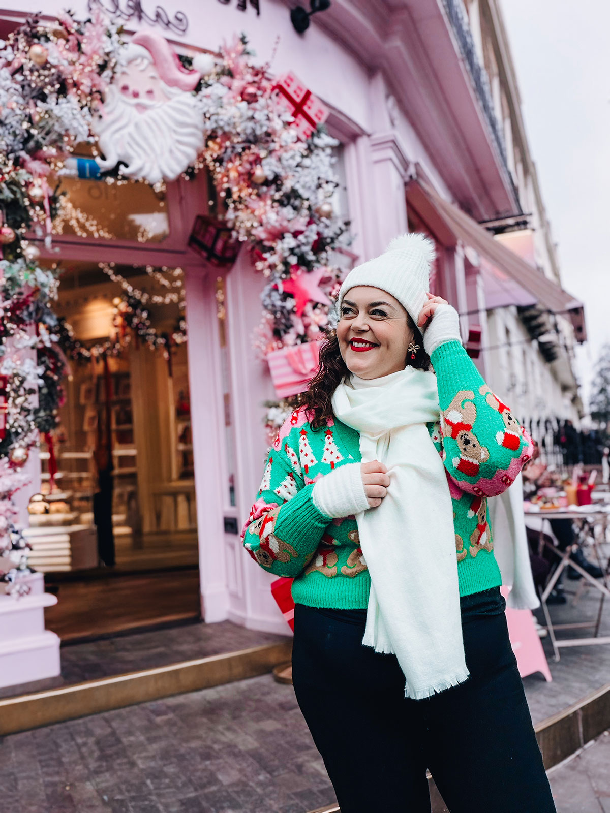 Rosie Parsons, professional photographer and business blogger, smiling in front of a pink decorated Christmas shop with a Santa face and frosted garlands. She wears a green Christmas jumper with festive print, white bobble hat and white scarf. The scene shows a creative christmas photoshoot location worth finding.