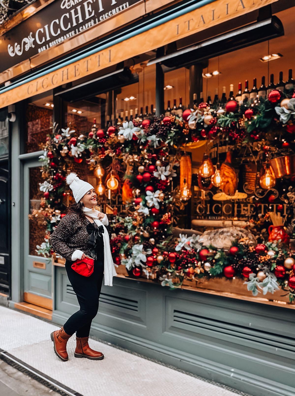Rosie Parsons, brand photographer and business blogger, smiling in front of a festive Christmas shop window display at Cicchetti restaurant. She wears a sparkly jacket, white bobble hat, white scarf, red bag and tan boots. The window features red baubles, greenery, warm Edison bulbs and berries, showing a vintage christmas photoshoot location.
