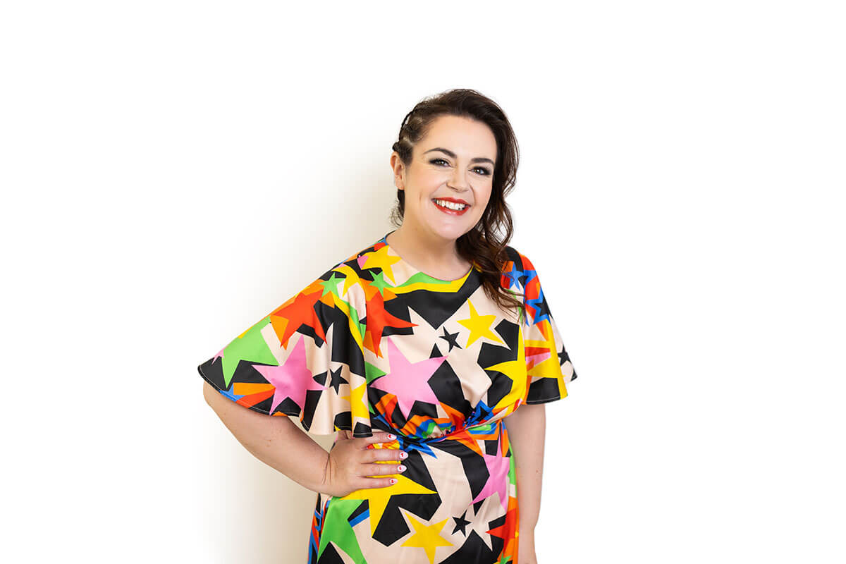 Smiling woman in a colourful, star-patterned fitted dress stands confidently against a plain white background. Her structured outfit shows what to wear for headshots when balancing bold style with flattering fit, reinforcing how clean lines photograph better than oversized clothing.