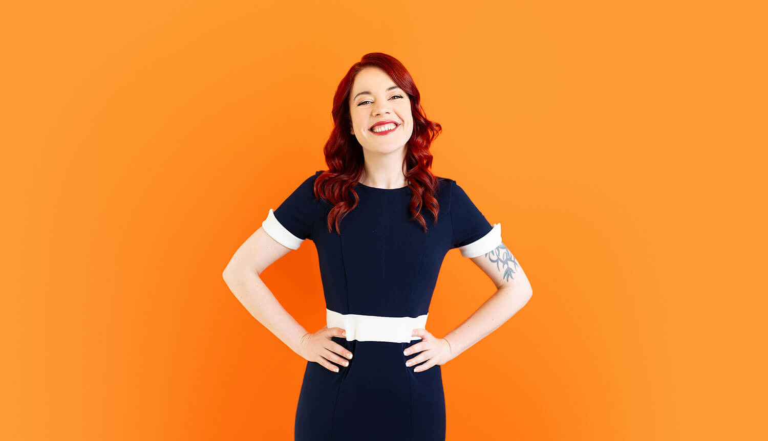 Smiling woman with red hair stands confidently in a navy dress with white accents, set against a bold orange backdrop. Her outfit shows how dark colours can retain detail and shape when paired with a high-contrast background and thoughtful lighting, avoiding the flattening effect of black on camera.