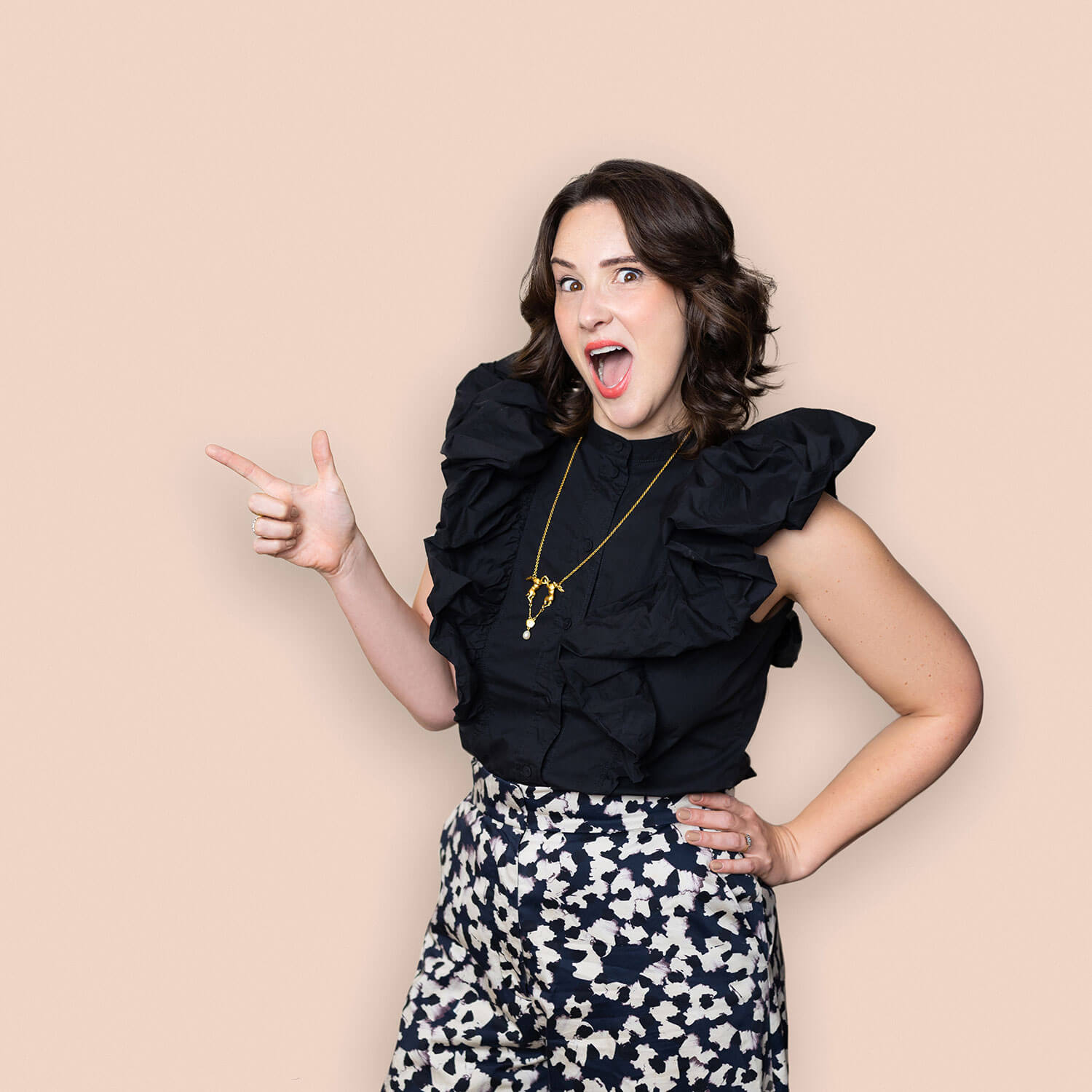 Woman striking a playful pose in a structured black ruffle top and patterned trousers, standing against a soft beige background. Her matte fabrics and confident styling highlight how thoughtful outfit choices—especially under studio lighting—affect the final headshot result.