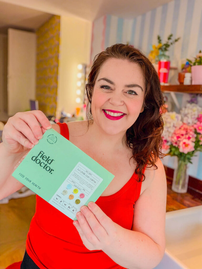 Rosie Parsons, brand photographer and business blogger, smiling and holding a Field Doctor healthy meal kit box. She wears a bright red top with her dark hair down, standing in a colourful room with pink flowers and shelves in the background. An image representing selfcare ideas, self care at home and making healthy lifestyle choices.