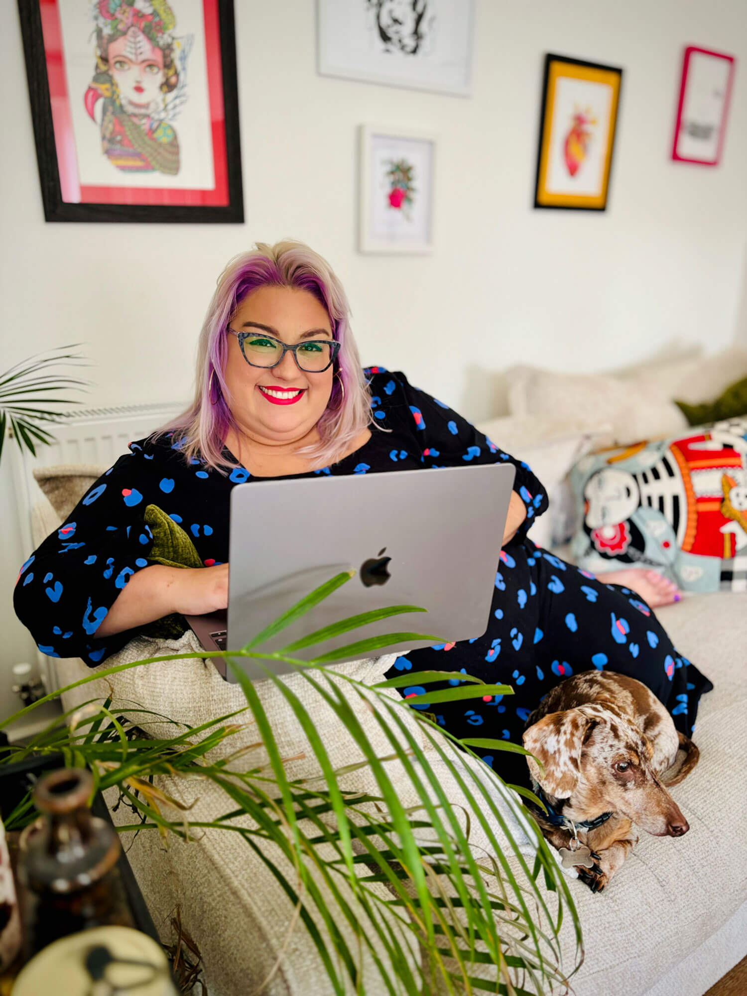 A woman with purple hair and glasses working on her blog with a MacBook on a cream sofa, wearing a black and blue leopard print dress with a cute dachshund beside her.