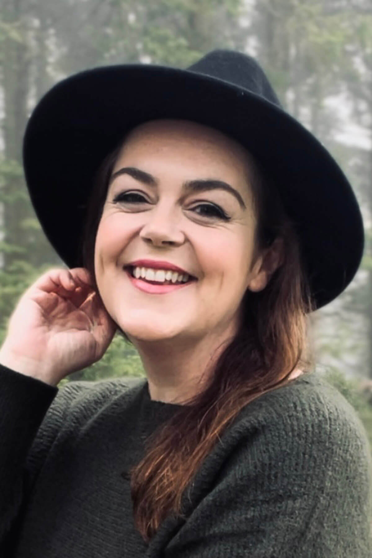 Woman smiling naturally outdoors wearing black fedora hat and grey sweater, hand gently touching hair near ear, with soft misty forest background – demonstrating relaxed aesthetic selfie pose with flattering natural light