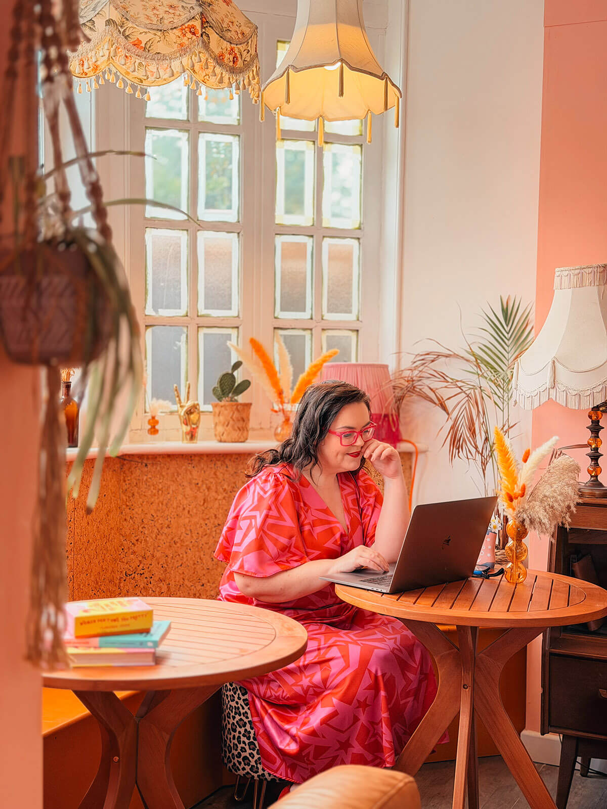 Rosie Parsons sits working on her blog on a laptop in a pink and orange styled room, wearing a star-print dress and pink glasses surrounded by vintage lamps and pampas grass