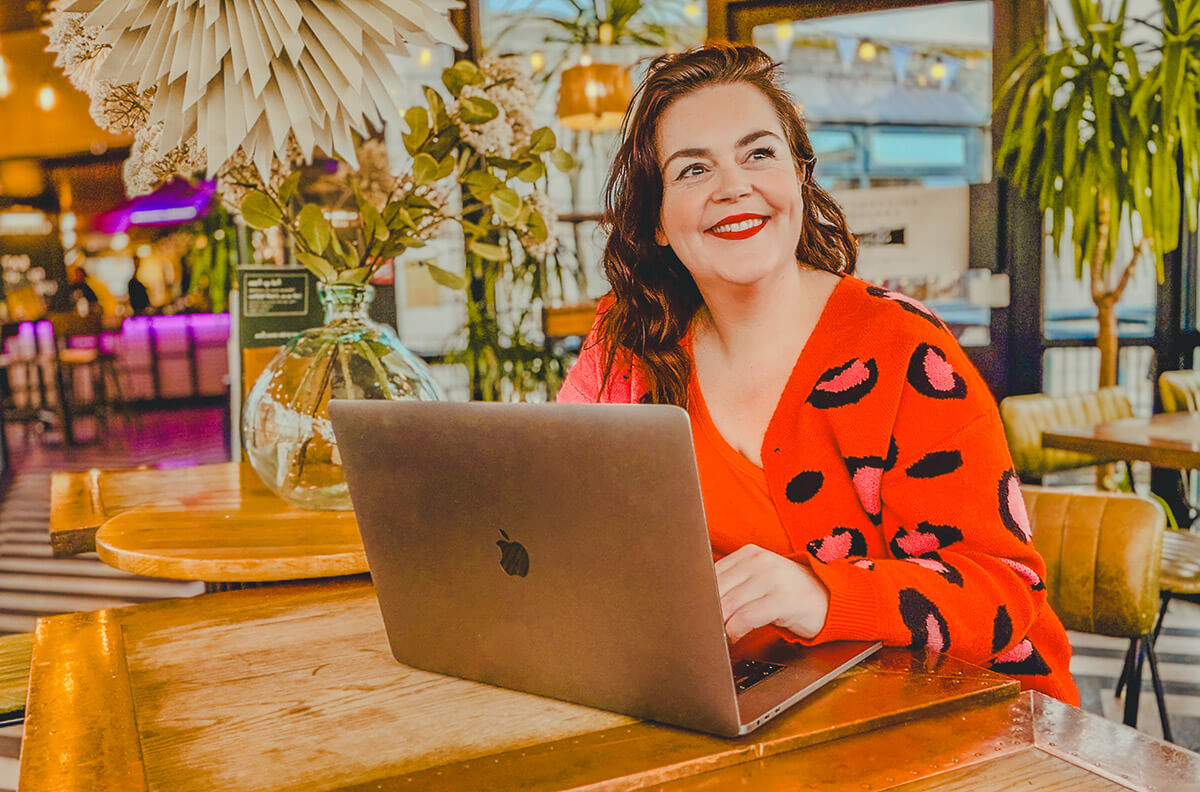 Blogger Rosie Parsons working on a MacBook in a stylish café, wearing a bold orange leopard print cardigan illustrating how to start a blog for beginners.