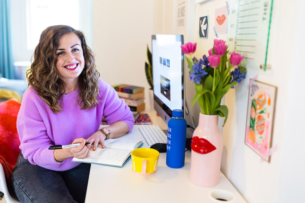 A woman in a lilac jumper planning in a notebook at a white desk with an iMac, fresh tulips, and colourful decor illustrating how to start a blog for beginners.