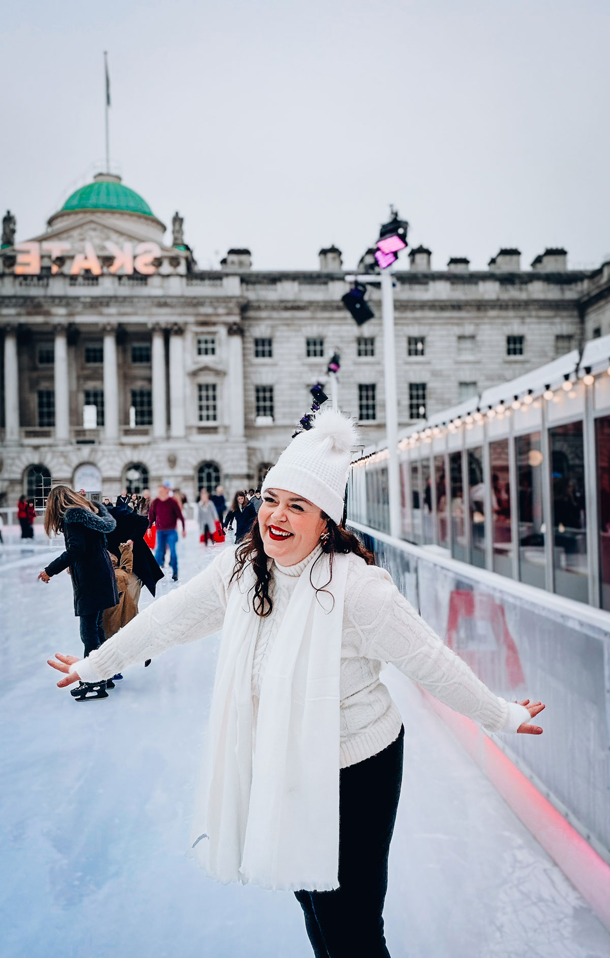 Rosie Parsons, headshot photographer and business blogger, smiling while ice skating at Somerset House in London. She wears an all-white outfit with a cream cable-knit sweater, white bobble hat and white scarf. The historic building is visible behind her, demonstrating christmas fashion photography with winter styling.
