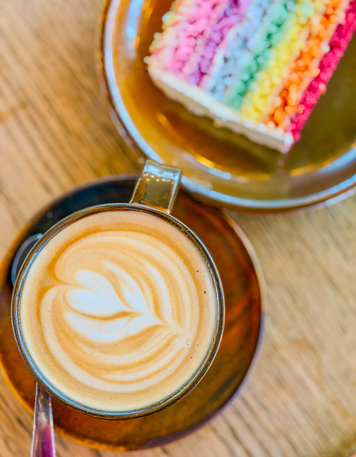 A flatlay of a latte with heart shaped latte art in an amber cup and saucer, next to a slice of colourful rainbow cake on a wooden table. A cheerful, indulgent image representing self care day treats, selfcare ideas and me time moments as part of a healthy lifestyle.