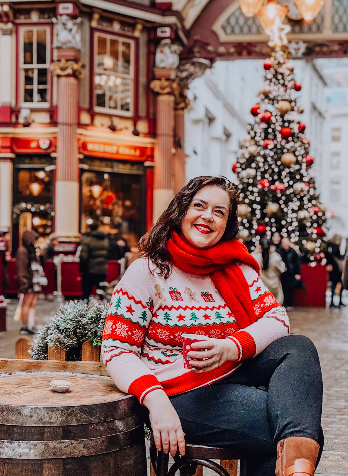 Rosie Parsons, professional photographer and business blogger, smiling while holding a hot drink at Leadenhall Market in London. She wears a festive fair isle Christmas jumper with trees and gingerbread print and a red scarf. A decorated Christmas tree with red and gold baubles glows behind her, showing a vintage christmas photoshoot location.