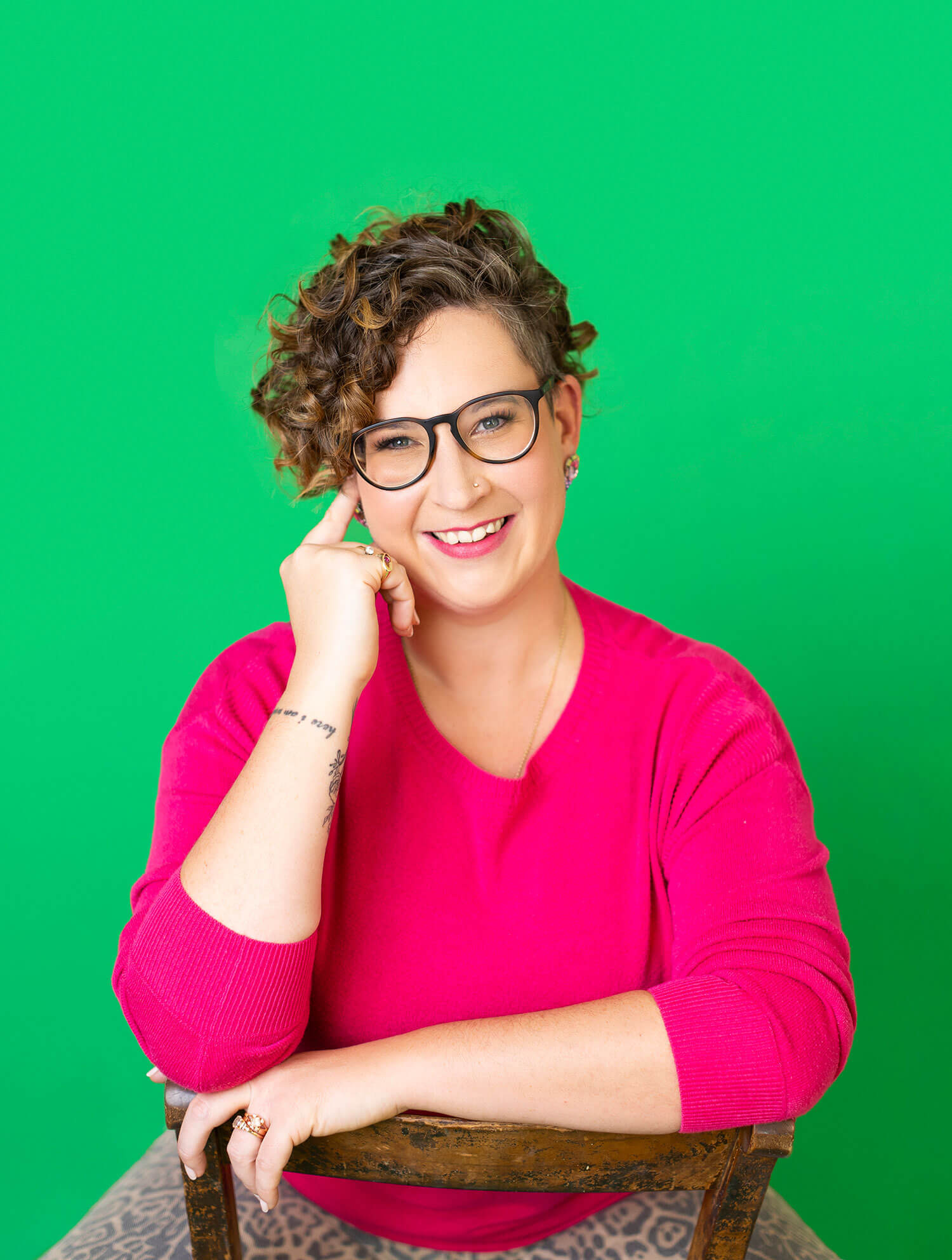 Smiling woman with short curly hair and glasses poses in a bright pink top against a vivid green background, seated confidently with her arm resting on a chair. Her high-contrast outfit and approachable expression demonstrate how bold colors and clarity make a LinkedIn profile photo stand out and feel memorable.
