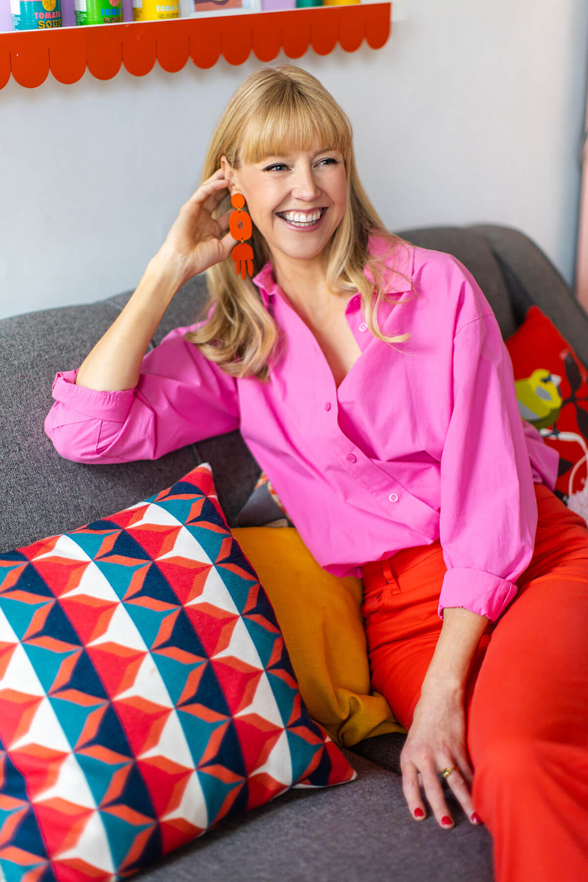 Personal branding photograph of a female service-based business owner smiling in a relaxed, informal setting. She smiles wearing bright pink shirt and orange statement earrings on colorful sofa, showing fun branding photoshoot inspiration for service-based business owners.