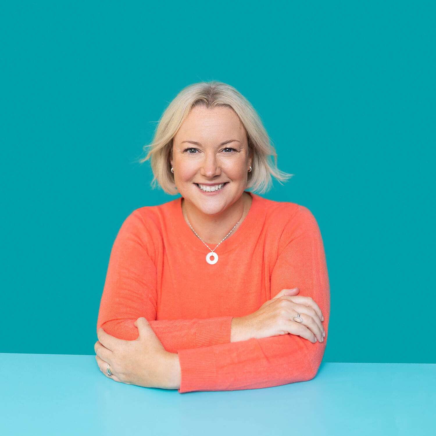 Smiling woman in a bright coral sweater sits with arms folded in front of a teal background, showcasing a clean, confident headshot with high contrast and minimal distractions. Her outfit draws attention to her face, supporting the message that headshot clothing should enhance rather than compete.